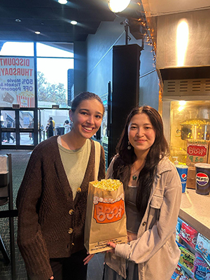 two students in lobby of theater, one holds bag of popcorn