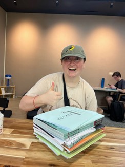 person sitting in front of stack of books, gives the thumbs up sign